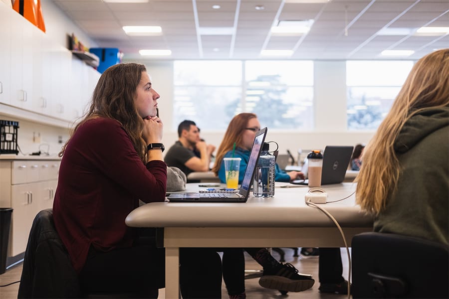 A student in a maroon top sits at a table with a laptop in front of her. There are other students around her in the classroom.