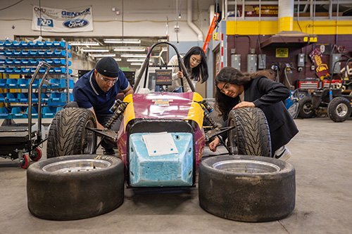 Three students work on a Formula-style race car in an engineering workshop.