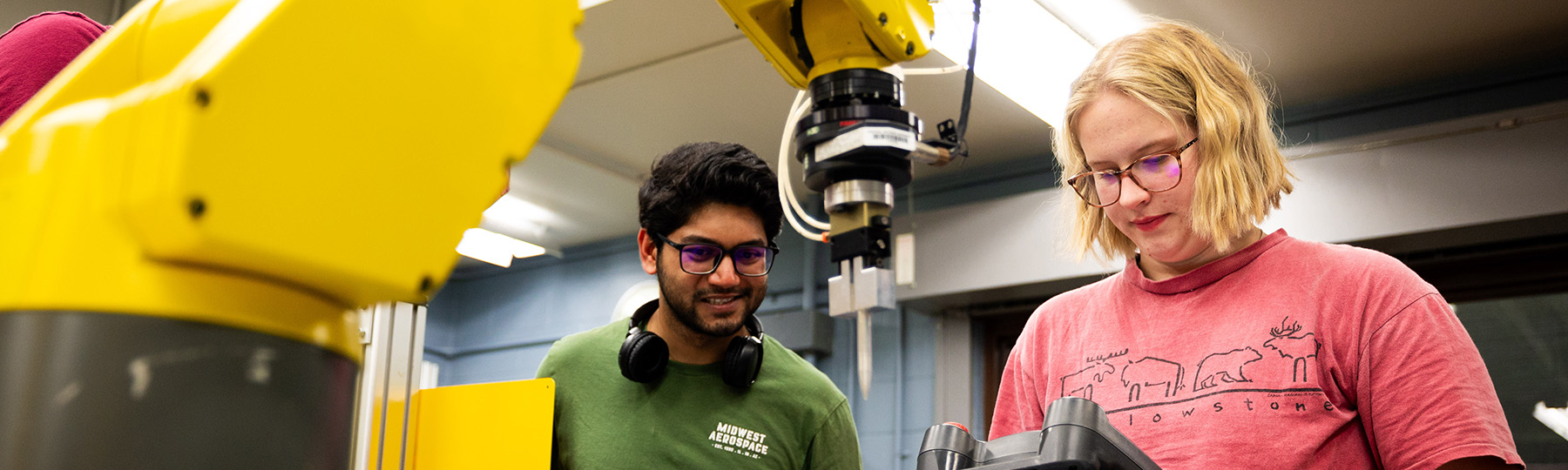 A man wearing headphones and glasses standing next to a machine while a woman with blonde hair holds the remote for a robotic arm.