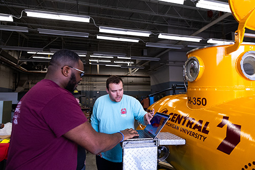 Two students programming life support settings on a laptop in front of CMU's one man submarine.