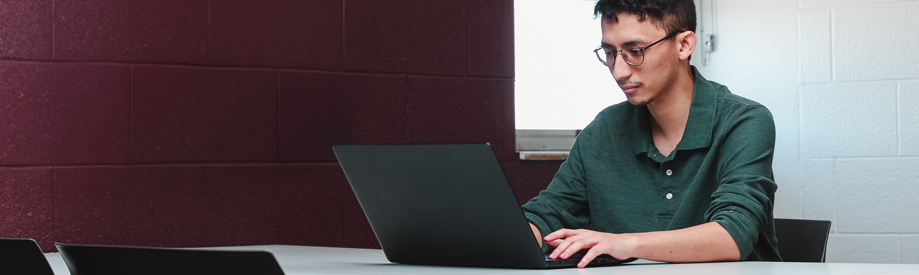 A student wearing glasses and a green polo shirt works on a laptop at a desk in a classroom setting.