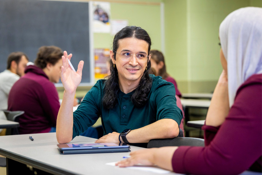 A student with long dark hair, glasses and an earring raises their hand while smiling during a discussion in a classroom. Another student in a headscarf listens across the table. Other students work in the background.
