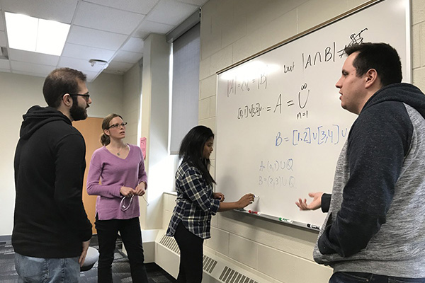 A group of students having a discussion in front of a white board with a mathematical equation on it.