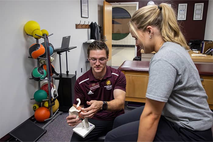An athletic training student sitting in the Injury Care Center holds an anatomical model and talks with a patient.