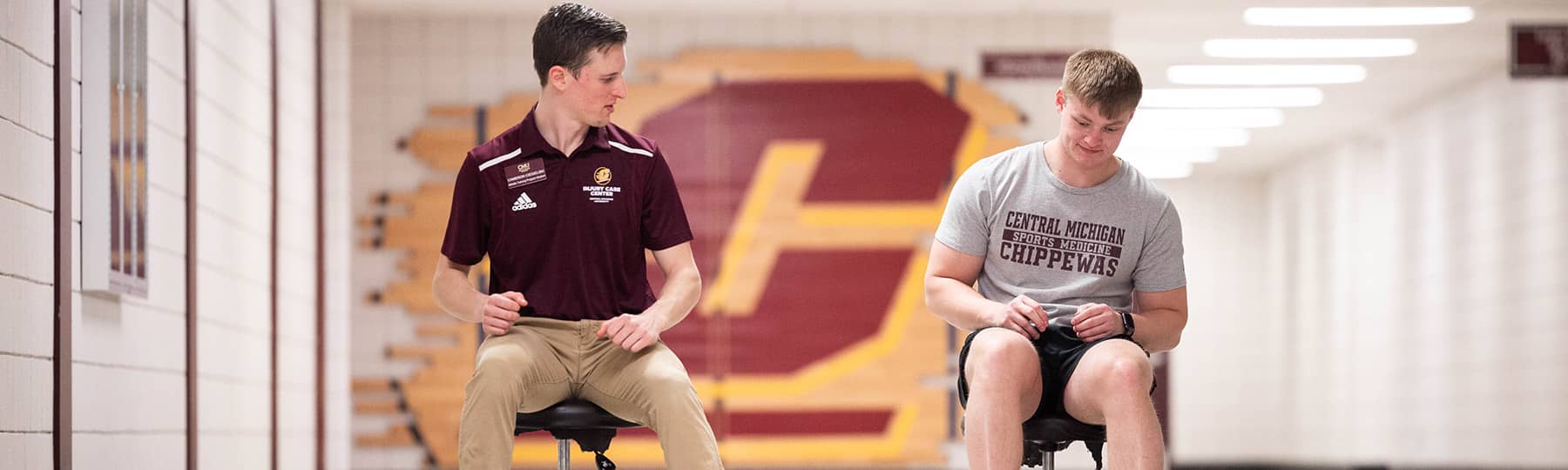 An athletic training student wearing a name tag, maroon polo, and khaki pants sits on a stool looking at a patient in a gray t-shirt sitting on a stool in the hallway outside of the Injury Care Center.