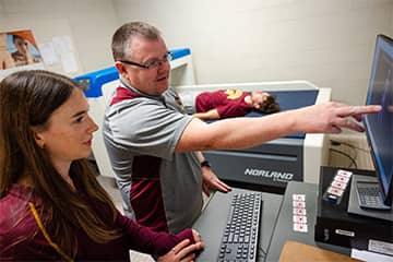 A student in a maroon t-shirt lying on a flat blue cushion undergoes a scan in the College of Health Professions with Dr. Paul O'Connor in a gray polo and an exercise science student in a maroon shirt examining a computer screen nearby.