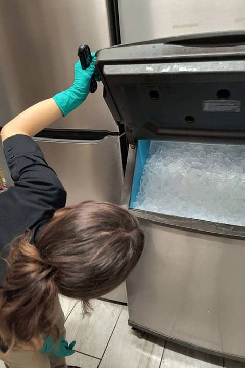 Linday Uzarski wearing a black shirt and teal colored gloves stands holding a black door of a stainless steel ice machine open. She leans over to look inside the ice machine.