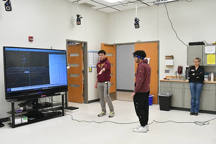 Motion Analysis Center 720x480 A student in a maroon sweatshirt directs another student who is standing front of a television screen with sensors placed on his sweatshirt and pants. Dr. Ustinova stands watching the students with her arms folded.