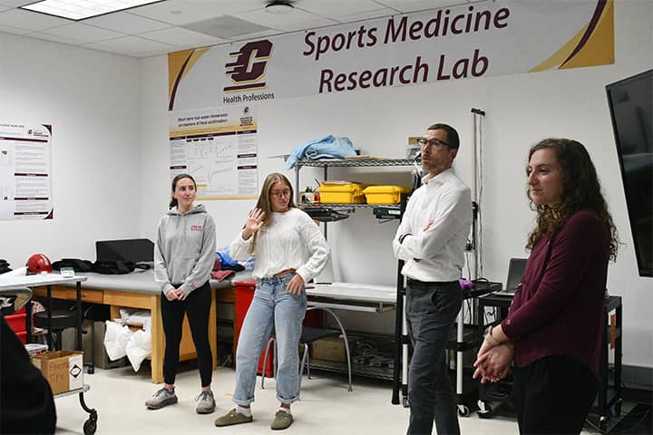 Sports Medicine Research Lab 720x480 Two students stand to the right and one to the left of Dr. Zuhl in the Sports Medicine Lab in the College of Health Professions. Research posters and banners are fixed to the wall and supplies are positioned behind them.