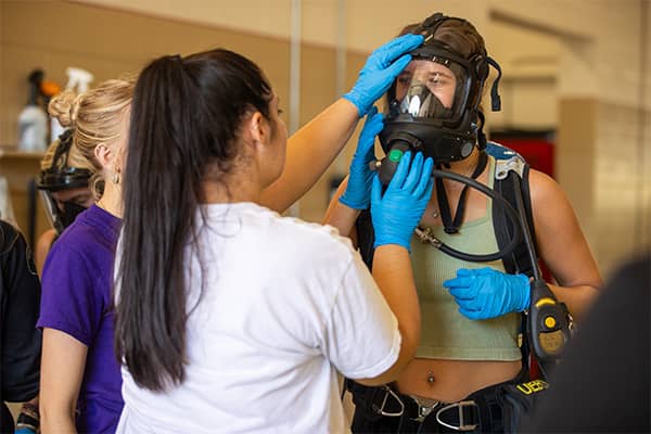 Hazmat Practice 600x400 A student with her hair in a pony tail wearing a white t-shirt and blue gloves holds a full-face respirator in place on her partner who is wearing blue gloves and an olive green tank top.