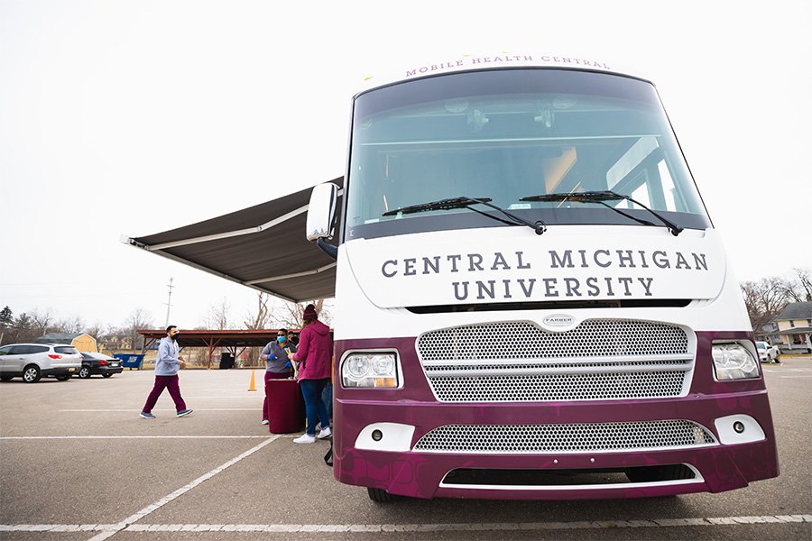 The white Mobile Health Central bus with Central Michigan University painted in maroon on the front is parked with its maroon awning extended. Three individuals are standing outside of the bus in the parking lot.