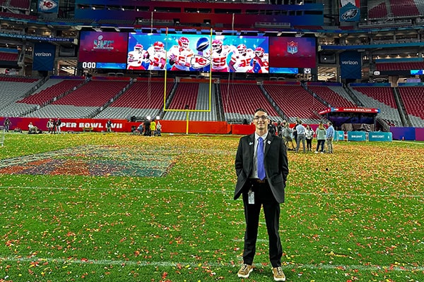 Sport management student, Quintin Hunsinger, stands on the State Farm Stadium football field covered in red and yellow confetti with the Super Bowl 57 Champions, Kansas City Chiefs football players pictured on the jumbotron behind him in the endzone.