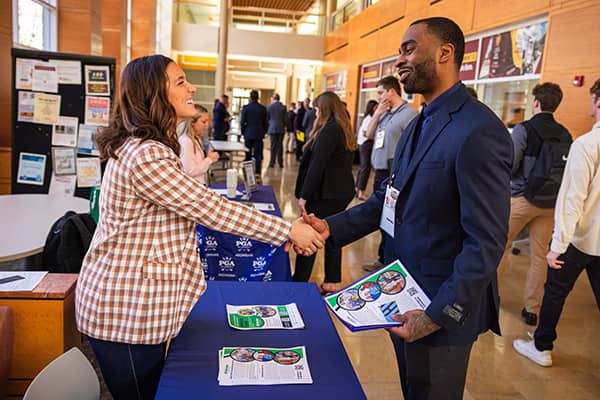 A woman dressed in a brown and white checkered blazer reaches across a blue table to shake hands with a man dress in a navy blue suit. The man is holding papers in his left hand.