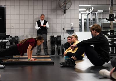 A student in a maroon t-shirt demonstrates a push-up position while three other students sit on the floor and watch in a weight room. Dr. Rick Ferkel stands in the distance evaluating the demonstration.