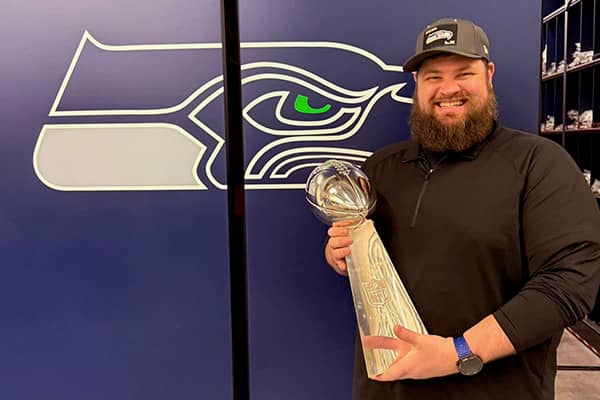 Lee Herteg, wearing a black quarter zip sweatshirt and a gray Seattle Seahawks baseball cap stands holding the Lombardi trophy in front of a Seattle Seahawks logo painted on a blue wall.