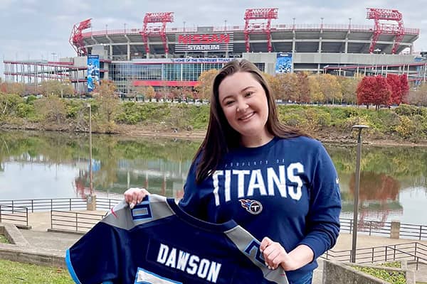 Taylor Dawson wearing a Tennessee Titans blue long sleeve shirt stands in front of a river outside of Nissan Stadium holding a Tennessee Titans football jersey with the name Dawson stitched across the back.