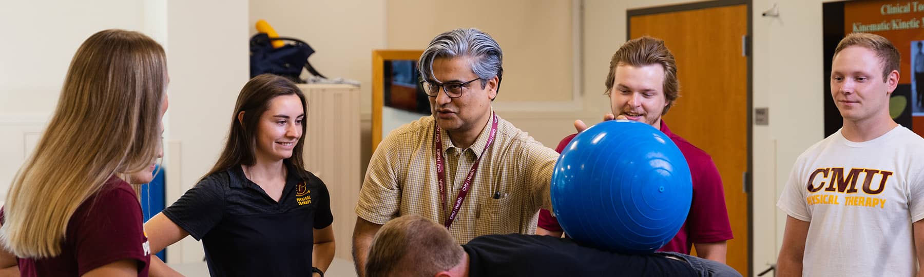 Dr. Raju Chowdhary, wearing glasses and a tan polo, stands and talks to four Physical Therapy students while holding a bright blue exercise ball on an individual's back with his left hand.