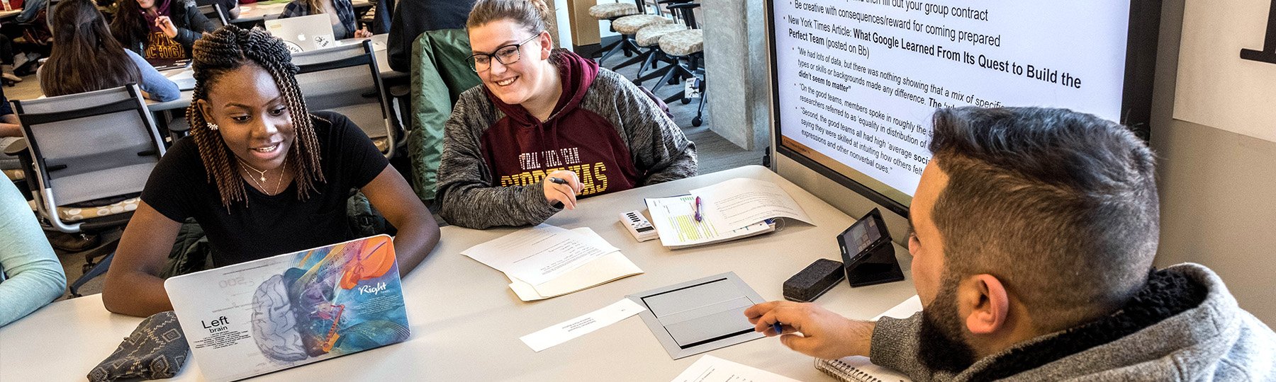 Three students sit around a table working on a laptop together.