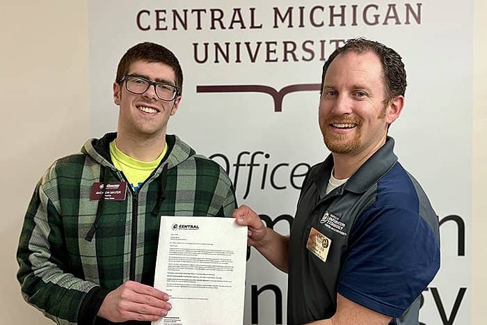 Andrew (left) is presented with his scholarship award letter by Derek (right). The two are standing in front of a wall with one of the CMU logos on it.
