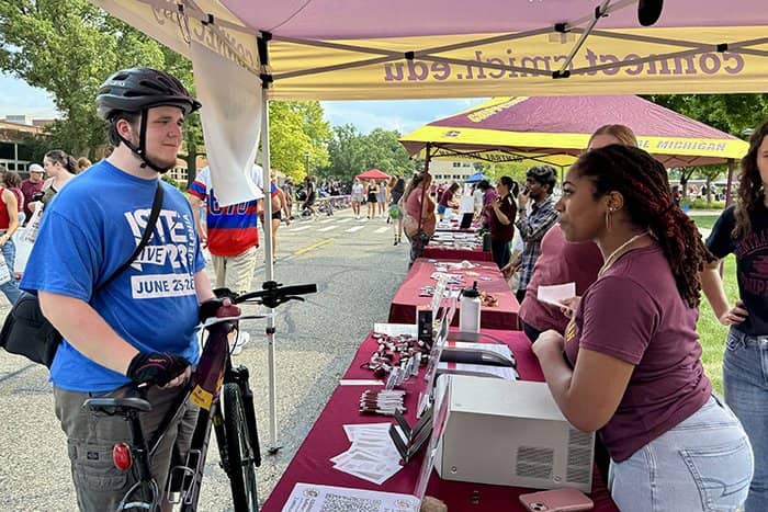 An OIT staff member talks with a student under a tent. The student is standing next to a bike and wearing a bike helmet and a blue shirt.