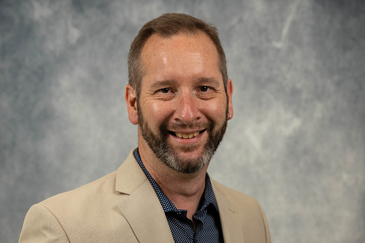 Portrait of Brian Roberts wearing a tan jacket over a blue shirt with a small white pattern on a marble gray background.