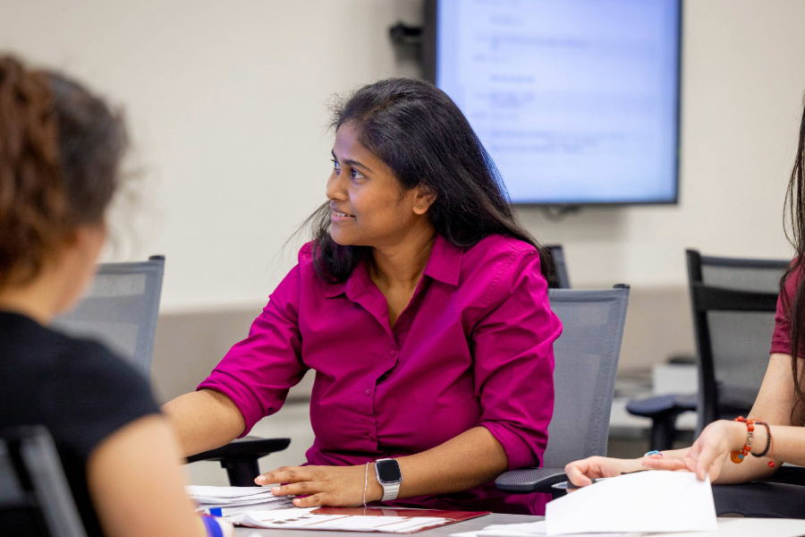 Student in a magenta shirt smiles while speaking with others during a classroom discussion. She sits at a table with papers in front of her, surrounded by others in a collaborative learning environment.