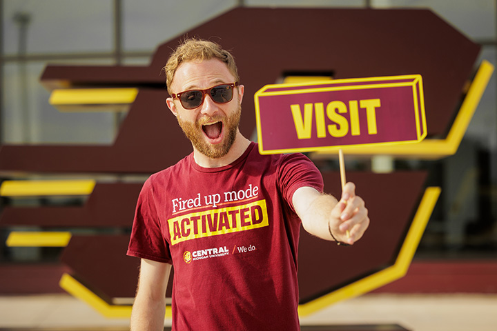 A male in his early 20s wearing sunglasses holds a small sign that says Visit.