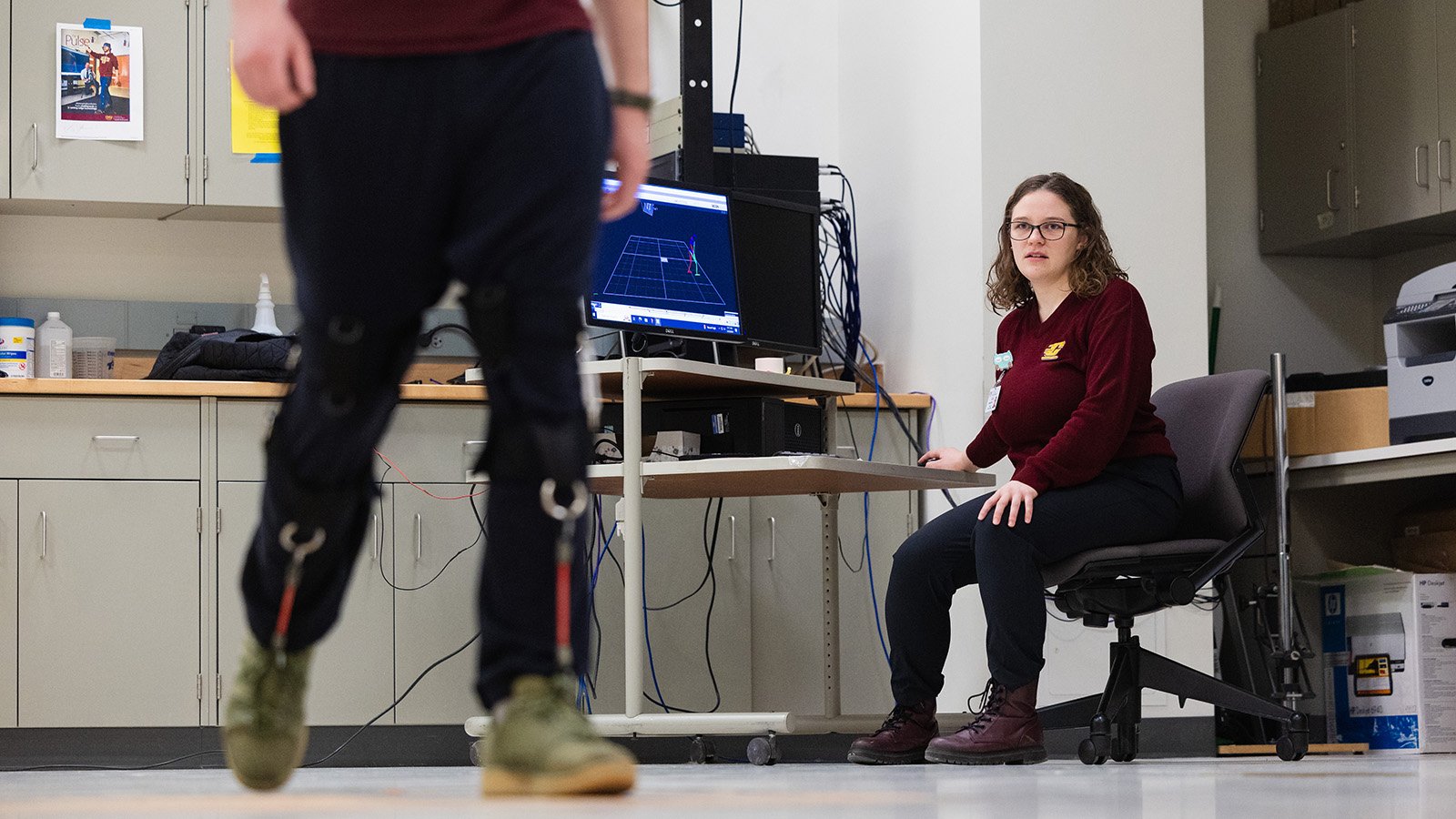 A physiology student sits in a chair in front of a computer as another student walks in the foreground wearing tracking devices.
