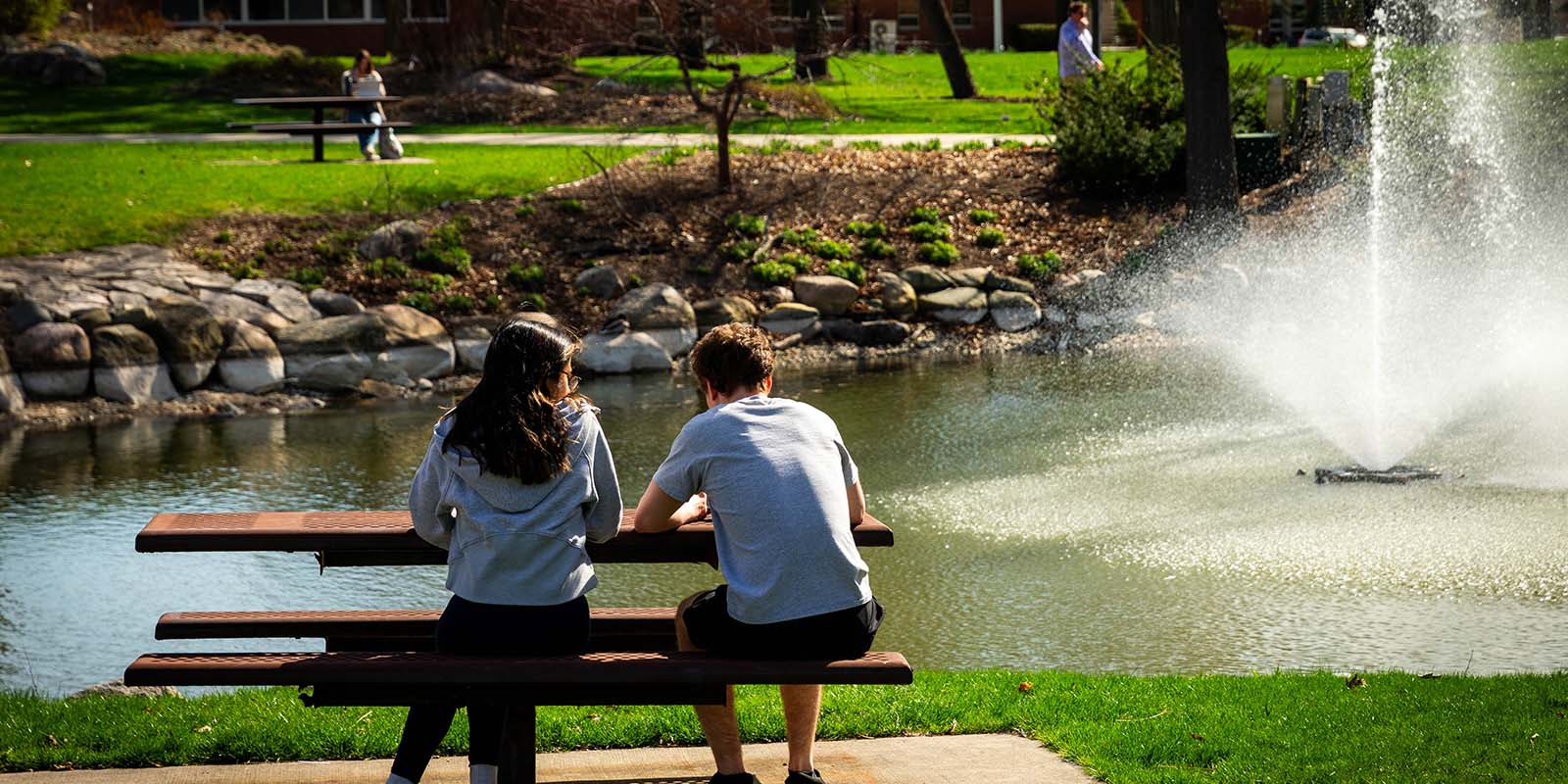 It's springtime on campus. a male and female student sit together at a picnic table by a landscaped pond with a central water fountain.