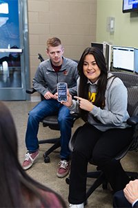 A woman holding a phone next to a man sitting in a chair in a computer lab.