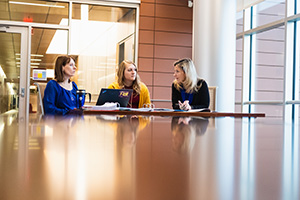 a group of women sitting at a table looking at each other and having a conversation with an open laptop and papers in front of them
