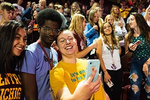 A group of people taking a selfie together while smiling with a group of people in the background behind them.