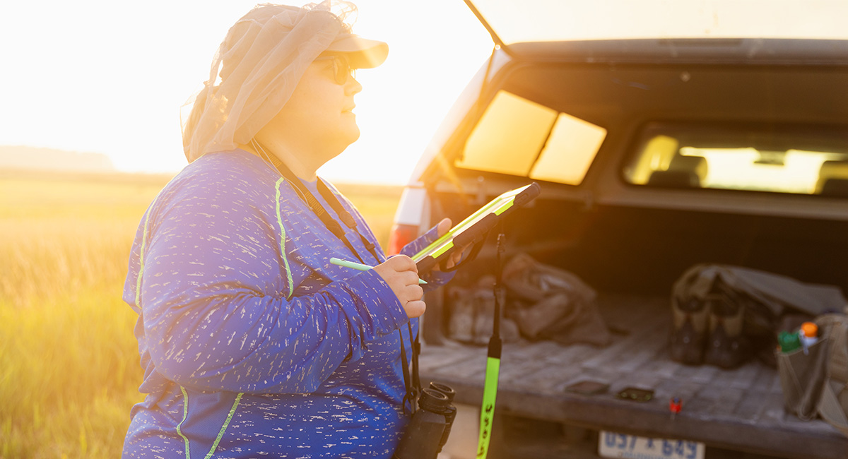 A student in a blue shirt, sunglasses and a netting hat, holds a clipboard standing behind the bed of a truck.