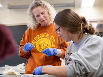 A woman in an orange shirt wearing blue latex gloves works with a student wearing glasses and blue gloves.