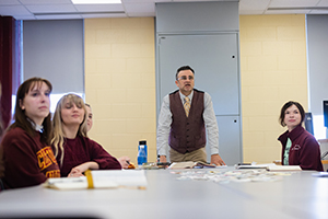 a man standing in front of a group of people sitting at a table while they stare past the camera observing something