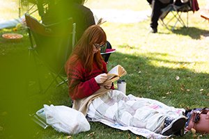 a woman sitting on the grass looking down and reading a book with a blanket over her lap