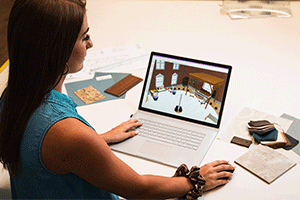 A fashion student works on a laptop while sitting at a table, reviewing color swatches and home decor.