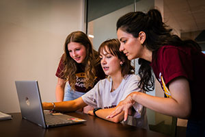 Three young women look at a laptop screen.