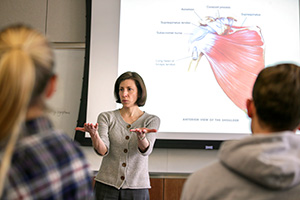 a woman standing in front of a projection screen that is showing muscles in the body and she is gesturing to the classroom as she teaches