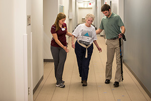 A man and women walk alongside the patient on both sides giving her assistance as they walk down a hallway