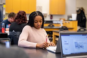 A female student sits at her desk looking down at her notebook with a pencil in her hand taking notes