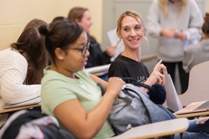 Two women sitting in a classroom at desks, one is smiling at the other while she pulls items out of her backpack.