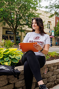 A young woman with long dark hair wearing a gray T-shirt sits on a low brick wall while balancing a laptop on her knee.