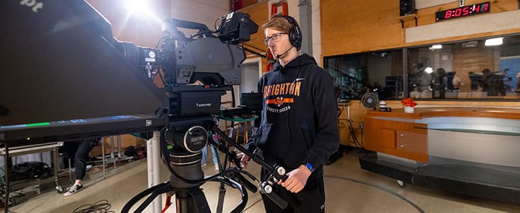 A young man wearing a headset stands behind a tv camera recording a performance.