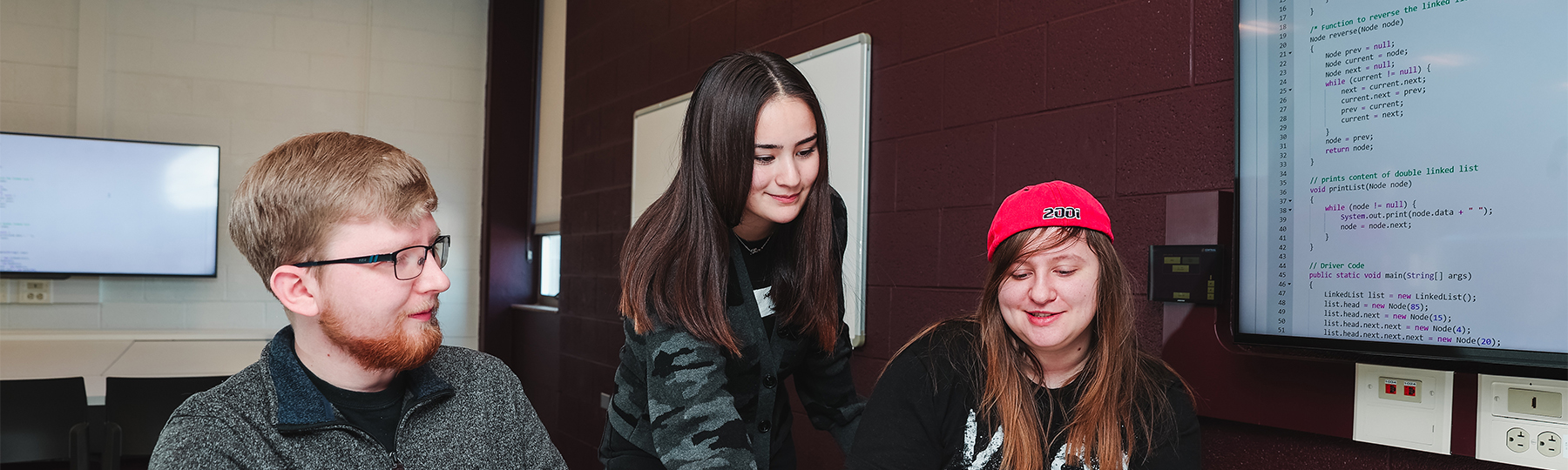 Three computer science students looking at a laptop and projecting code onto a monitor in a study space.