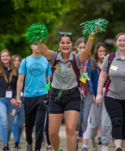 A CMU orientation leader waves green pompons and smiles at the camera while leading new students across campus.