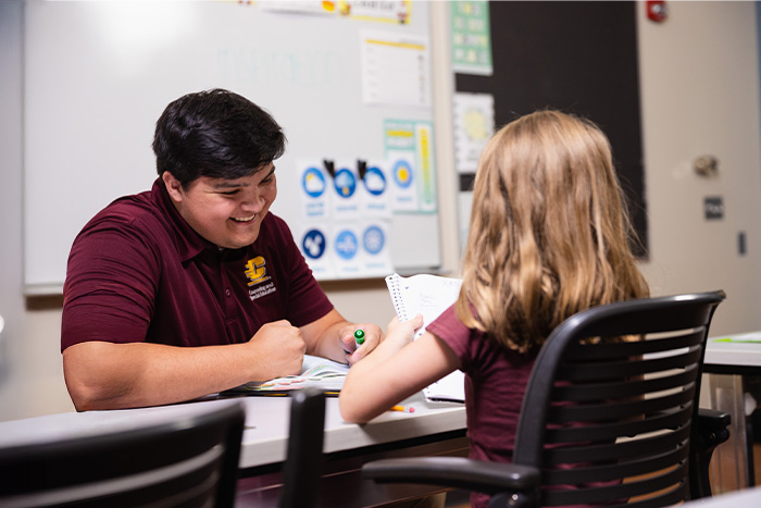 A student tutor sits at a table helping a young child with a workbook in a classroom.