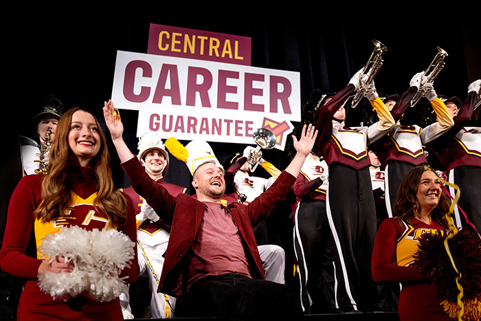 A man seated on a stage raises his arms while the CMU marching band and cheerleaders stand next to him and behind him holding a large “Central Career Guarantee” sign.