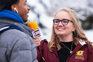 A CMU student interviews another student outside on a cold winter day, while they both have winter jackets on and the interiewer holds a maroon and gold microphone up to the student being interviewed.