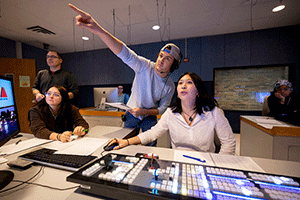 Siz students in a sound studio working during a recording session, sitting near a sound board. One student is pointing and looking into the distance.